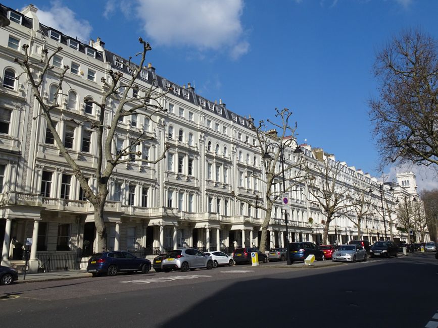 A street scene showing a row of traditional brick and white-painted residential buildings with large sash windows, some featuring decorative cornices and brickwork detailing, situated along a busy urb